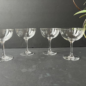 May include: Four clear champagne coupe glasses with etched starburst designs. The glasses have a wide, shallow bowl and a slender stem, perfect for serving celebratory drinks. The glasses are arranged on a dark surface.