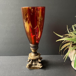 May include: An antique, tall, red-orange glass vase with a decorative brass base. The vase has a flared shape and sits on a detailed, ornate base. The glass has a reflective, glossy finish. A houseplant is in the background.