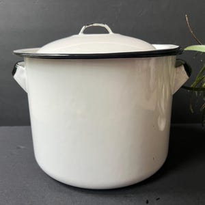 May include: A white enamel stockpot with a matching lid, featuring a black rim around the top edge and the lid. The pot has two side handles and a smooth, glossy finish, presenting a cylindrical shape with a flat base and a domed lid top.