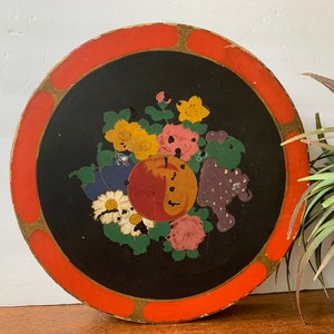 May include: A round wooden tray with a black center and a red and gold rim. The center features a painted design of flowers, fruit, and leaves.