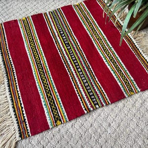 May include: Red and white striped rug with black and yellow accents. The rug has a fringe border and is made of woven wool.