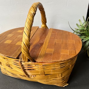 May include: A vintage wicker picnic basket with a wooden lid and a woven handle. The basket is made of natural brown wicker and has a rectangular shape.