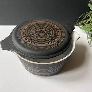 May include: A black and white bowl with a matching lid. The lid features a brown spiral design. The bowl has a white interior and a black exterior with horizontal lines. The bowl has two handles.