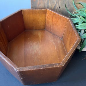 May include: A vintage, octagonal wooden bowl with a warm brown finish. The bowl's interior and exterior showcase the natural wood grain. The design features a wide, open interior, perfect for display or storage. The bowl is set against a neutral background.