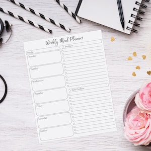May include: A white Weekly Meal Planner with sections for each day of the week, produce, and non-produce. The planner is on a white wooden surface, with a pen, notebook, and decorative items in the background.