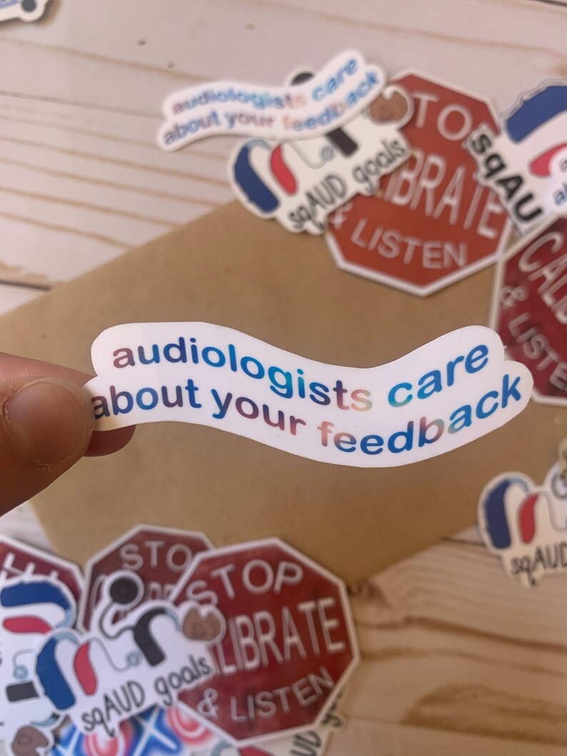 Op de afbeelding: Een set kleurrijke stickers met de tekst "audiologists care about your feedback" en "STOP & LISTEN" op een rode stopbordachtergrond. De stickers bevinden zich op een bruin houten oppervlak.