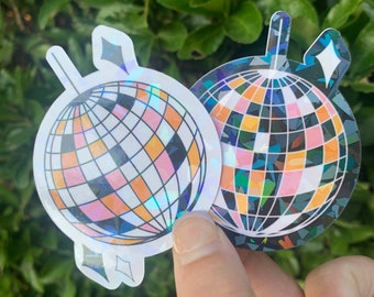 May include: Two iridescent disco ball stickers. One sticker has a black and white design with a rainbow iridescent finish. The other sticker has a pink, orange, and white design with a rainbow iridescent finish.