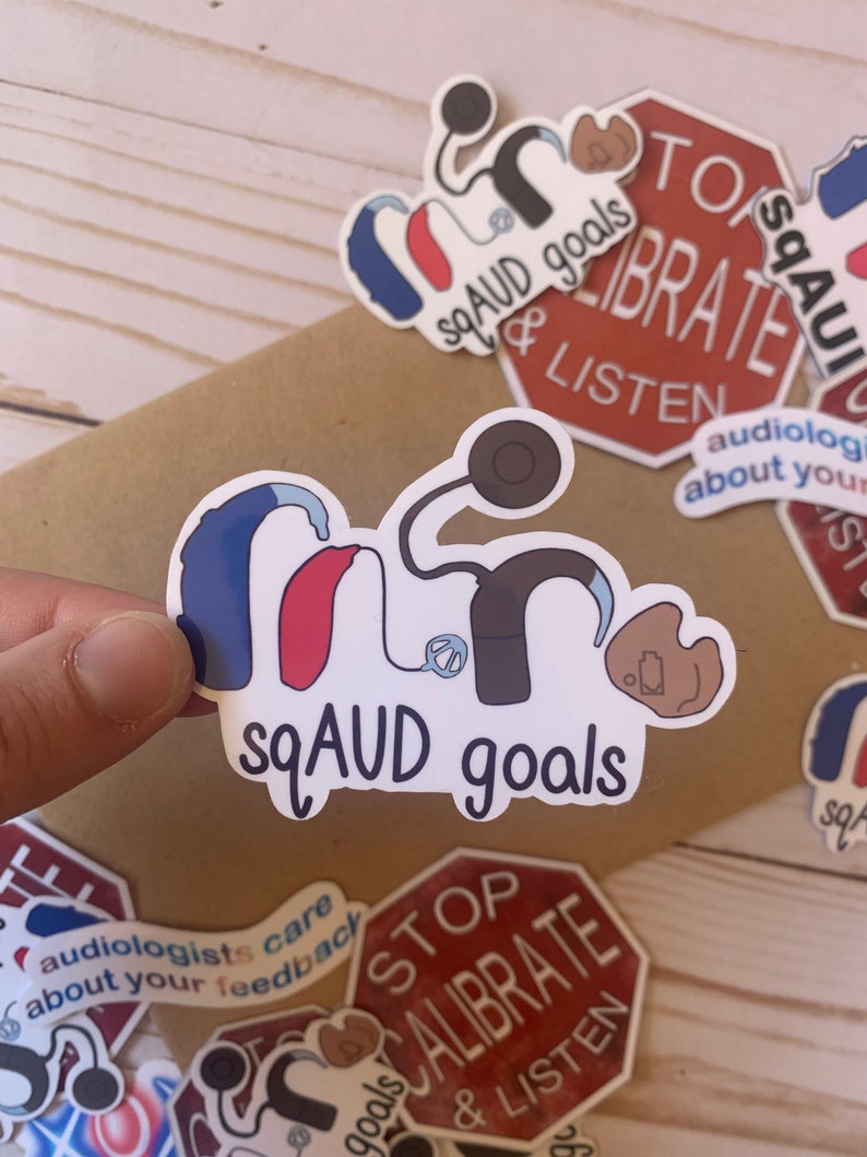 Op de afbeelding: Een set stickers met een gehoorapparaat, een stethoscoop en de tekst "sqAUD goals". De stickers bevinden zich op een bruin houten oppervlak.