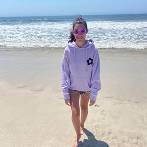 May include: A person wearing a lavender hoodie with a black flower graphic on the front, standing on a sandy beach with the ocean in the background.