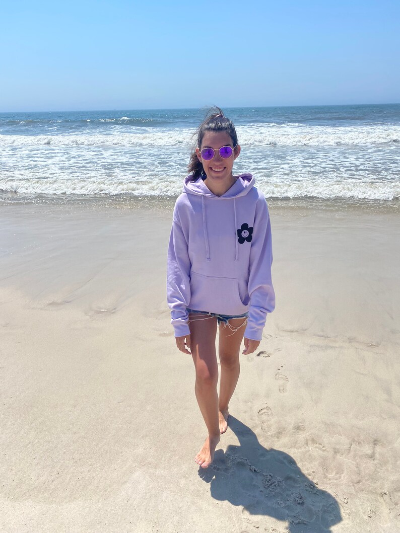 May include: A person wearing a lavender hoodie with a black flower graphic on the front, standing on a sandy beach with the ocean in the background.