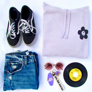 May include: A flat lay of a light purple hoodie with a black flower with a smiley face on it, a pair of black canvas shoes, a pair of blue jeans, a pair of pink sunglasses, a black vinyl record, and a keychain with a purple tag that says "Stay Groovy" and a white tag that says "Love that journey".