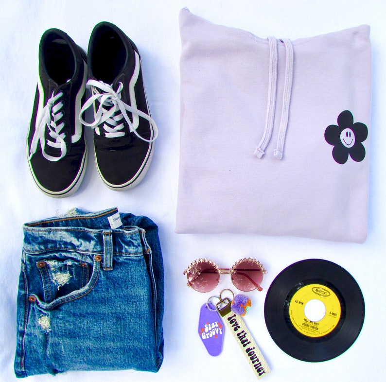 May include: A flat lay of a light purple hoodie with a black flower with a smiley face on it, a pair of black canvas shoes, a pair of blue jeans, a pair of pink sunglasses, a black vinyl record, and a keychain with a purple tag that says "Stay Groovy" and a white tag that says "Love that journey".