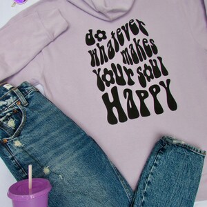May include: A lavender hoodie with a black graphic that says "do whatever makes your soul happy" in a retro font. The hoodie is paired with blue jeans and a purple cup.
