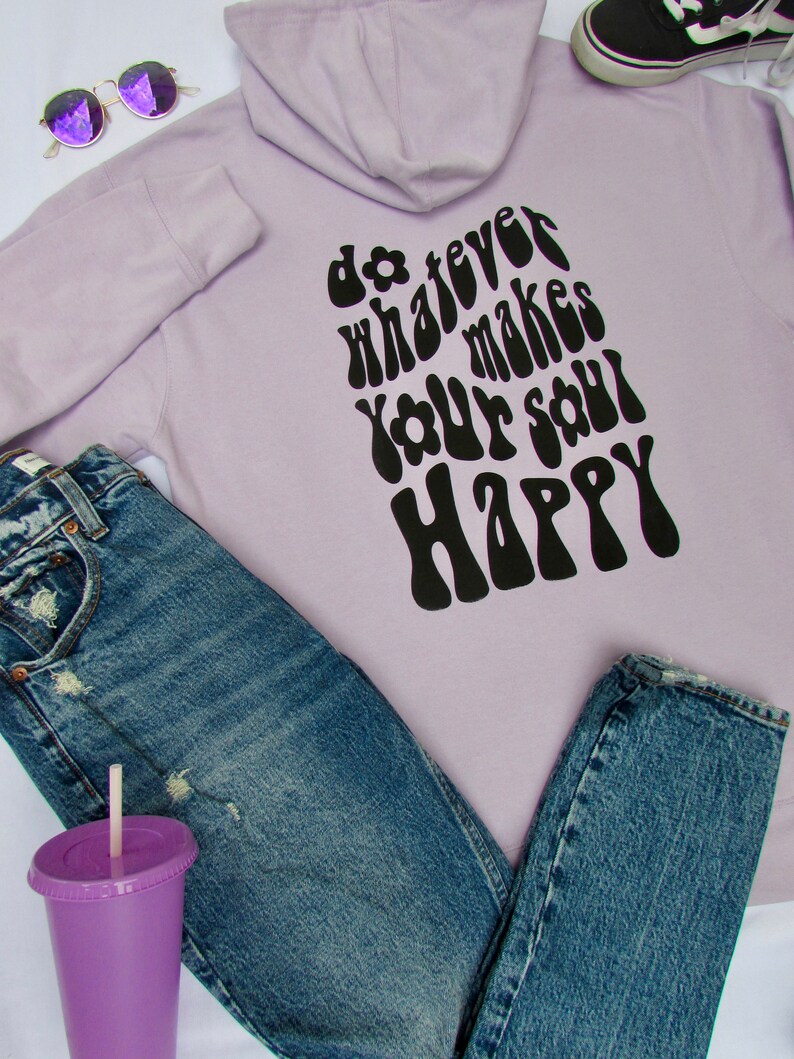 May include: A lavender hoodie with a black graphic that says "do whatever makes your soul happy" in a retro font. The hoodie is paired with blue jeans and a purple cup.