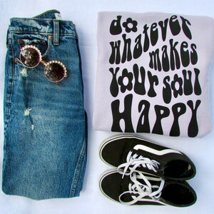 May include: A light purple sweatshirt with the text "do whatever makes your soul happy" in black, retro-style font. The sweatshirt is paired with blue jeans, black and white sneakers, and sunglasses.