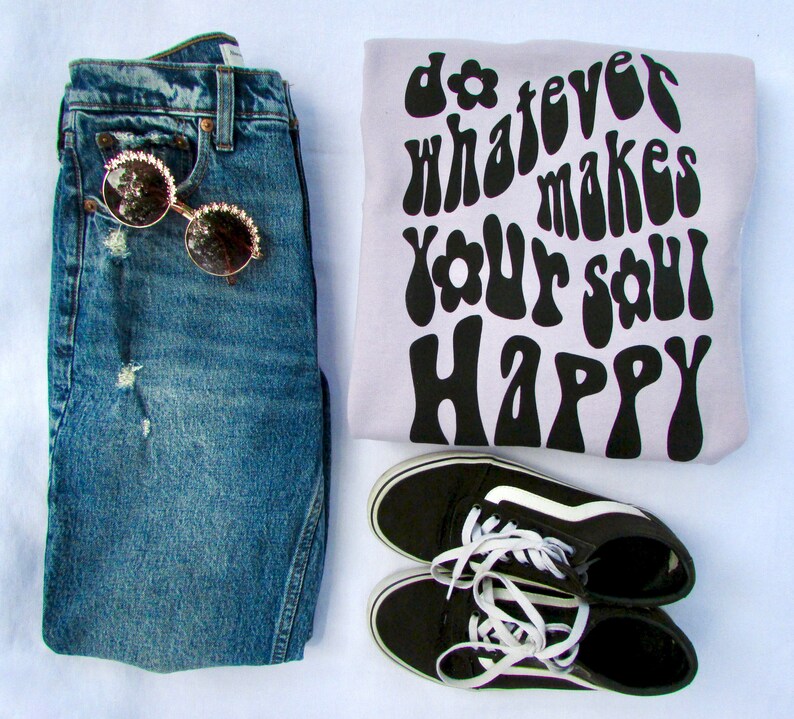 May include: A light purple sweatshirt with the text "do whatever makes your soul happy" in black, retro-style font. The sweatshirt is paired with blue jeans, black and white sneakers, and sunglasses.