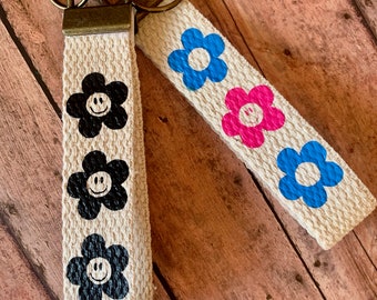 May include: Two keychains with a beige canvas strap and a metal ring. The straps have a repeating pattern of black, blue, and pink flowers with smiley faces.
