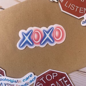 Op de afbeelding: Een witte sticker met het woord "XOXO" in blauwe en rode neonletters. De sticker zit op een bruin oppervlak.