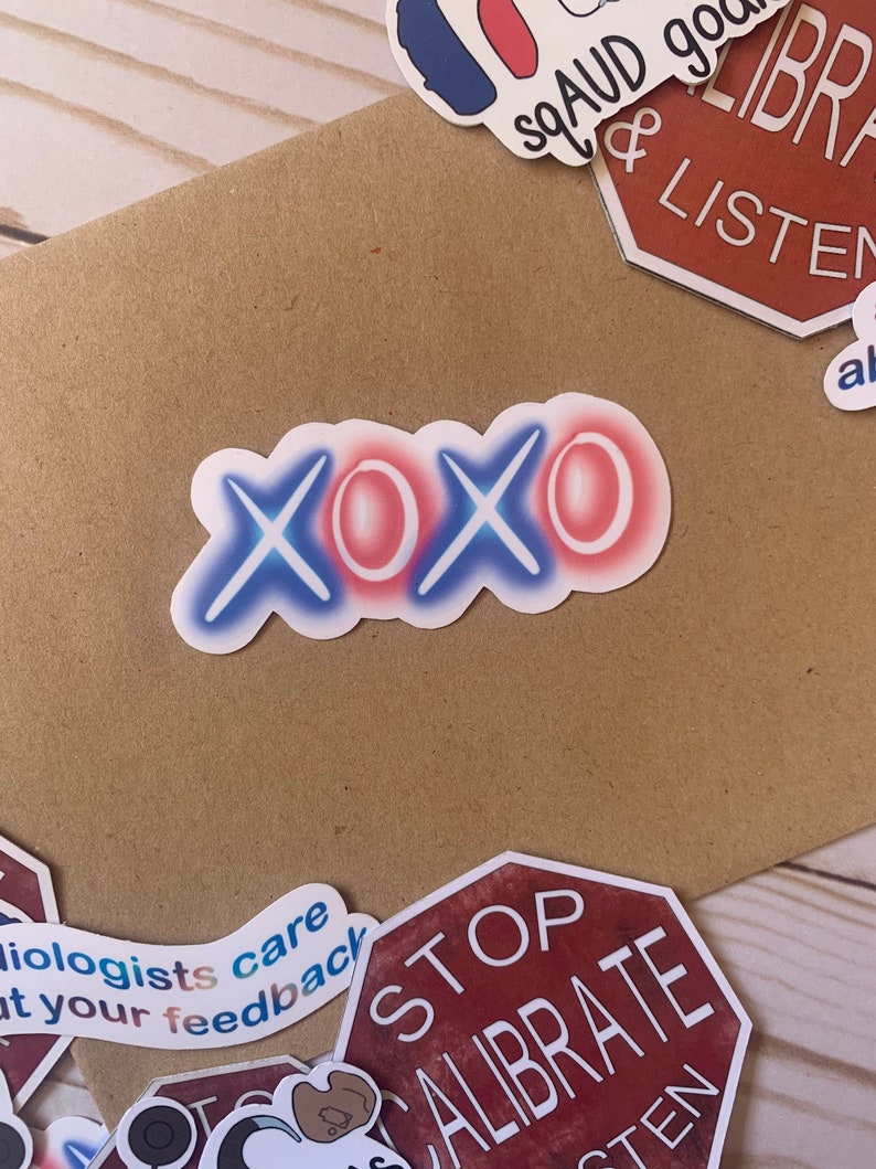 Op de afbeelding: Een witte sticker met het woord "XOXO" in blauwe en rode neonletters. De sticker zit op een bruin oppervlak.