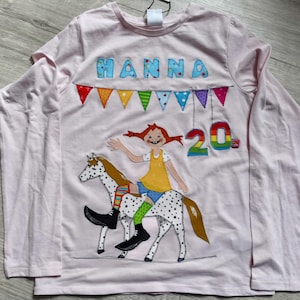 Pode incluir: Uma camisa rosa claro de manga comprida com um gráfico colorido. O gráfico apresenta o nome "HANNA" acima de uma faixa de bandeiras coloridas, uma menina montando um cavalo manchado e o número "20".