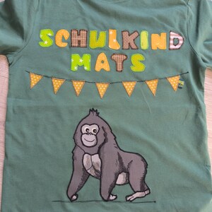 May include: Green short-sleeved t-shirt with the text "SCHULKIND MATS" in colorful patchwork letters above a cartoon gorilla. A string of yellow pennants with white polka dots is below the text.