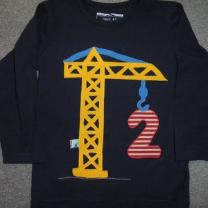 May include: A navy blue long-sleeved shirt with a yellow construction crane and a red and white striped number two. The crane has a blue hook and the shirt has the text "Next 87" on the front.