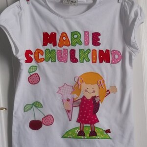 May include: White t-shirt with the text "Marie Schulkind" in colorful embroidered letters. The shirt also features an embroidered image of a girl holding a bouquet of flowers.