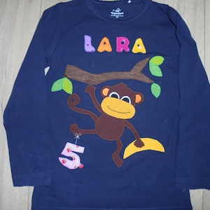 May include: A navy blue long-sleeved shirt with a monkey holding a banana and a number 5 balloon. The shirt has the name "LARA" in colorful letters.