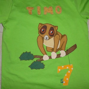 May include: Green t-shirt with a brown lemur sitting on a branch with green leaves. The lemur has large brown eyes and white paws. The number 7 is sewn on the shirt in orange with white stars. The name "TIMO" is sewn on the shirt in brown letters.