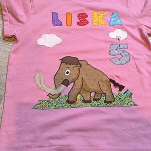 May include: Pink t-shirt with the name "LISKA" in colorful letters and a number "5" with a cloud. A brown mammoth with white tusks is featured on the front, standing on a patch of green grass.