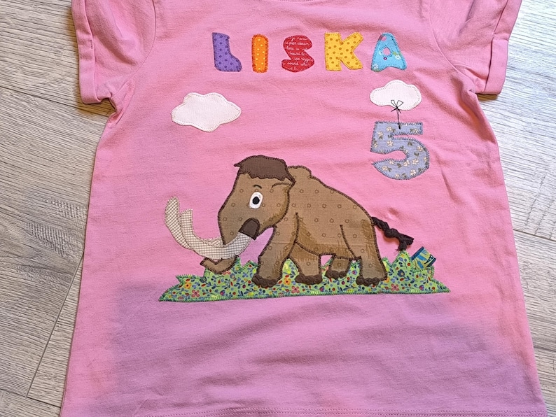 May include: Pink t-shirt with the name "LISKA" in colorful letters and a number "5" with a cloud. A brown mammoth with white tusks is featured on the front, standing on a patch of green grass.