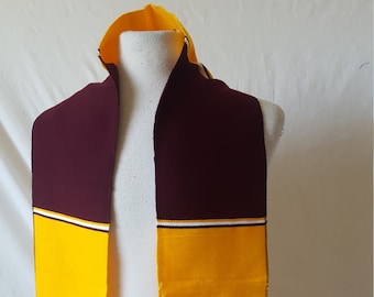 May include: A maroon and gold graduation stole with white stripes. The stole has fringe on the ends.
