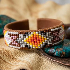 May include: A handcrafted cuff bracelet with a brown leather base and a beaded design. The beadwork features a geometric pattern in shades of white, pink, brown, gray, yellow, and orange. The bracelet is displayed on a textured surface with stones.