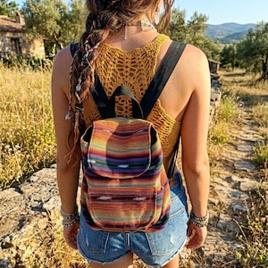 May include: A small backpack featuring a vibrant, multi-colored horizontal striped pattern in shades of orange, red, purple, and blue. The bag has a top flap closure, black shoulder straps, and a front pocket, worn over a yellow crochet top and denim shorts.
