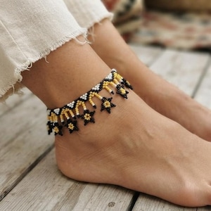 May include: An anklet crafted from small seed beads in a repeating pattern of black, white, and yellow. The design features a series of dangling star-shaped charms along the lower edge, creating a decorative fringe effect around the ankle.