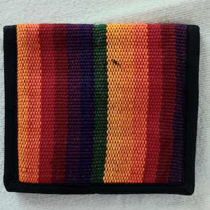 May include: A woven wallet with a vibrant rainbow striped pattern in shades of red, orange, yellow, green, and purple. The wallet is rectangular with a black border. The texture of the weave is visible.