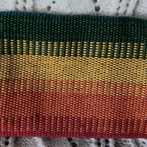 May include: A rectangular wallet with a woven design in green, yellow, orange, and red. The wallet has black edges and a textured appearance. The color gradient creates a vibrant and eye-catching accessory.