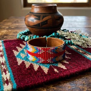 May include: A cuff bracelet featuring a geometric pattern of small, multi-colored glass beads in shades of red, blue, yellow, and white, set against a tan leather interior lining. The bracelet has an open-ended design with a visible gap.