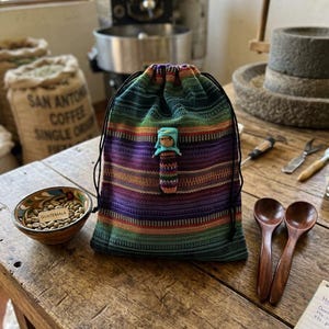 May include: A colorful, striped drawstring bag with a small doll attached. The bag is next to a bowl of coffee beans labeled "Guatemala" and two wooden spoons. The background includes coffee roasting equipment.