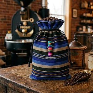 Puede incluir: Una bolsa de tela con cordón y rayas horizontales en tonos azul, verde y beige, llena de granos de café tostados. Una pequeña muñeca decorativa de colores está sujeta a la parte frontal de la bolsa, que descansa sobre una superficie de madera.