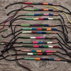 May include: A collection of twelve handmade friendship bracelets featuring multi-colored woven thread segments in various hues including red, green, blue, orange, purple, and white, each finished with long black cord ties for adjustable closure.