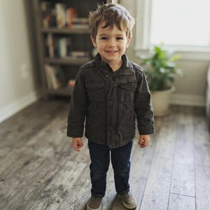 May include: A dark charcoal gray utility jacket with a stand-up collar, front zipper closure, and two chest pockets, paired with dark blue denim jeans and gray canvas sneakers. The jacket features a structured design with visible stitching details.