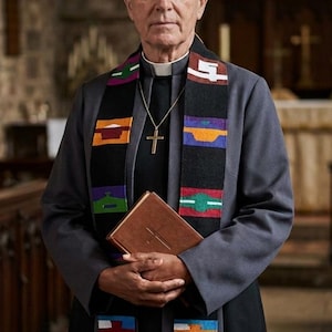 May include: An older man in a dark gray clerical suit, holding a brown book and wearing a colorful stole with geometric patterns. A gold cross hangs from his neck. The background is a church interior.