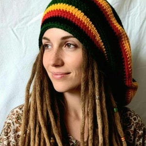 May include: A crocheted hat in the colors of green, yellow, and red. The hat is worn by a person with long, brown dreadlocks. The person is wearing a patterned top.
