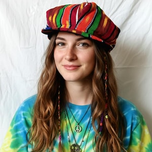 May include: A colorful, striped hat with a flat brim. The hat features a vibrant mix of red, orange, green, and yellow. The person is wearing a tie-dye shirt and multiple necklaces with peace symbols.