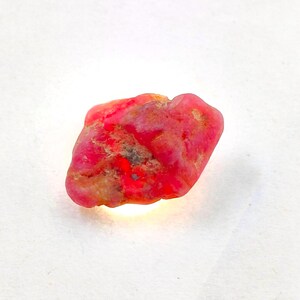 May include: A raw, unpolished gemstone with a vibrant, saturated pinkish-red hue. The stone features an irregular, organic shape with a textured, pitted surface and subtle variations in color intensity, including small areas of darker inclusions.
