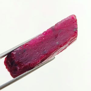 Peut inclure: Un brut de rubis non taillé, maintenu par une pince argentée. Le rubis est d'un rouge profond et saturé, avec une surface rugueuse et texturée. La pierre précieuse est de forme rectangulaire et semble fraîchement extraite.