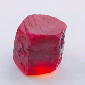 Pode incluir: Uma gema de rubi bruta e não cortada. O rubi é de uma cor vermelha profunda e saturada, com uma superfície áspera e texturizada. A gema tem uma forma retangular com a parte superior e inferior ligeiramente arredondadas. A parte inferior do rubi tem uma aparência brilhante e translúcida.