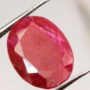 May include: An oval-cut, vibrant red gemstone with multiple facets that reflect light. The stone is held by a pair of metal tweezers, showcasing its translucent quality and deep crimson color, which displays subtle internal inclusions and variations.