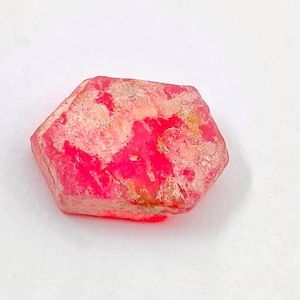 May include: A hexagonal-shaped, vibrant pink gemstone with a textured surface. The stone has areas of lighter pink and beige, creating a marbled effect. The gemstone is set against a white background.